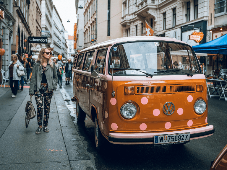 A woman walks next to an orange bus in a downtown street. Are you going to the next F1 race in Austria?